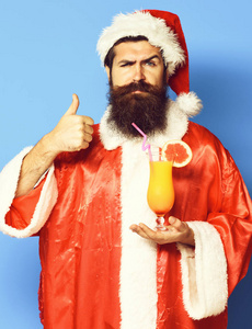 帅有胡子的圣诞老人人与非酒精性鸡尾酒杯举行圣诞节或圣诞毛衣和蓝色工作室底色上显出很酷的新的一年帽子的严重脸上长长的胡须