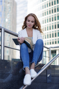 美丽的女人坐在台阶上，在摩天大楼之间看书