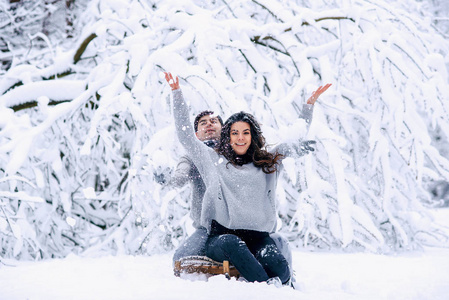 美丽的孕妇和她的丈夫坐在雪撬上，在一个下雪的冬天公园里，他们玩得很开心，吐着雪。 家庭爱和关怀的概念。