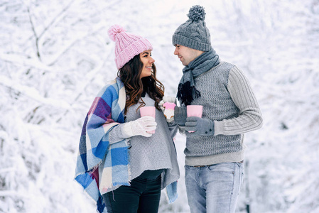 幸福的年轻夫妇走在一个雪地公园里，喝着粉红色的茶和粉红色的饼干。 穿着暖和的漂亮孕妇和她的丈夫。