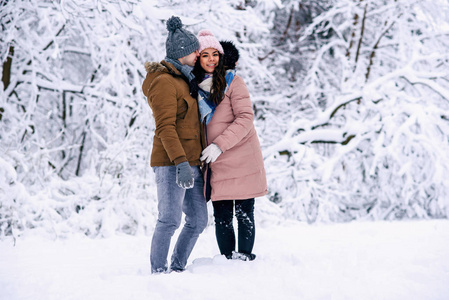 美丽的孕妇穿着温暖的衣服，和她可爱的丈夫一起在下雪的冬天公园散步。