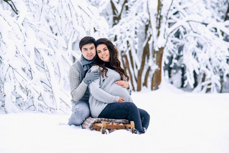 美丽的孕妇和她的丈夫坐在雪橇上在一个下雪的冬天公园。