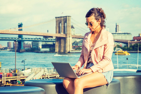 年轻的美国女人在纽约工作，穿着粉红色的皮夹克，蓝色的牛仔短裤太阳镜，坐在河边，用笔记本电脑工作。 布鲁克林曼哈顿桥的背景