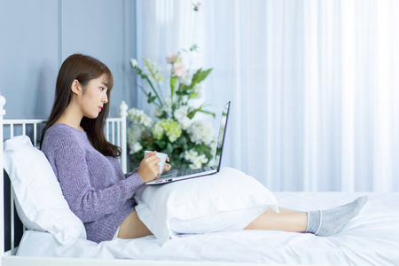 亚洲女人在床上工作。她穿毛衣紫罗兰色。早上检查电子邮件互联网从笔记本。
