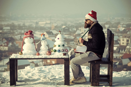 带铅笔和笔记本的嬉皮士坐在椅子上。 圣诞老人在冬日写作。 圣诞节和新年假期庆祝活动。 雪人和木制桌子上的圣诞树。 学习和有趣的概