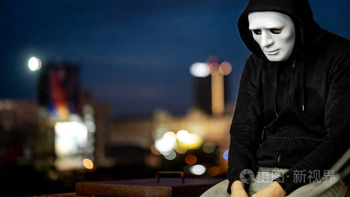 神秘的帽衫人戴着白色面具在黄昏时坐在废弃建筑的屋顶上双相情感障碍