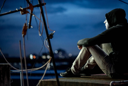 神秘的帽衫人戴着白色面具,在黄昏时坐在废弃建筑的屋顶上.