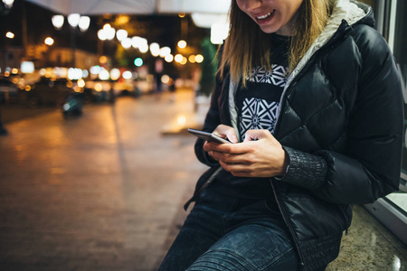 快乐的年轻女子在街上使用手机和微笑,低头看着发光的屏幕灯的夜晚
