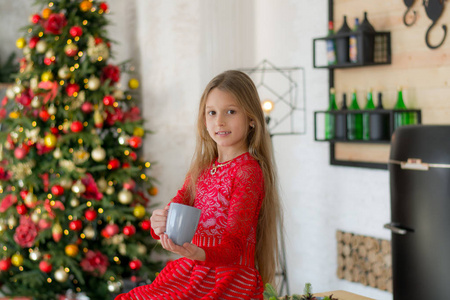 穿着红色裙子的漂亮女孩坐在小厨房里微笑。 杯子在孩子手里。 圣诞树附近的婴儿