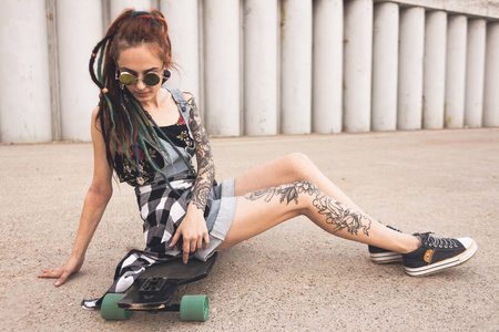 有纹身和长发绺的年轻女孩坐在长板在混凝土结构的背景下