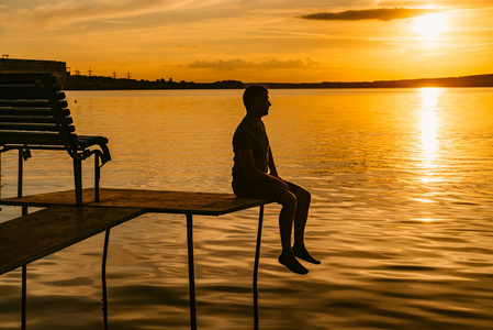 日落时独自坐在木桥上的人的轮廓。 孤独的人在一座小桥上看着明亮的夕阳反射在水中。