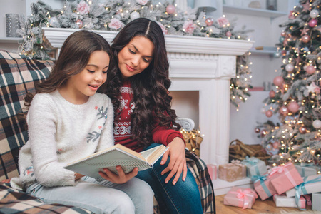 母亲和女儿坐在一起看书。他们看了。女孩用双手拿着它。它们在装饰过的房间里。他们身后有圣诞树和壁炉