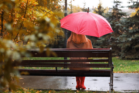 秋天公园里带着雨伞坐在长凳上的女人。 雨天