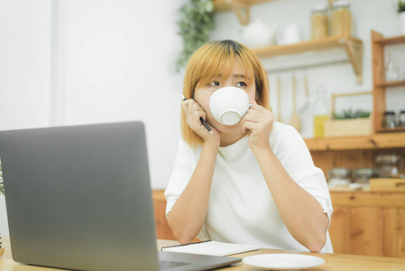 美丽的年轻微笑的女人在笔记本电脑上工作,一边享受着在家客厅里喝着