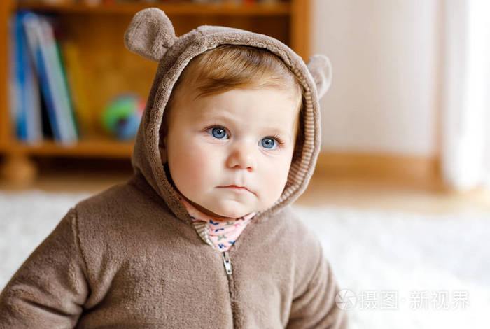 蓝眼睛的女婴穿着棕色熊冬天外形在阳光明媚的卧室。刚出生的孩子正穿着户外散步。儿童托儿所。为儿童提供纺织品和床上用品。幸福健康的女