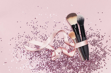不同的化妆品化妆刷与粉红色丝带和全息闪光纸屑的形式，星星在粉红色背景平躺顶部视图复制空间化妆配件假日生日新年。