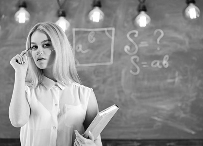 老师在眼镜上看起来很自信, 站在教室里, 黑板上的背景。带书的女人开始上课, 在摘眼镜时凝视观众。有吸引力的讲师概念