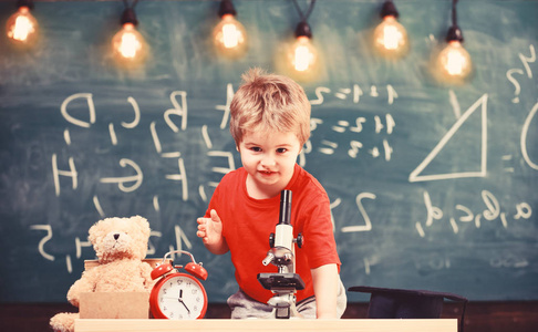 第一个对学习教育感兴趣的人。 小男孩在教室黑板上的显微镜附近。 孩子在钟和泰迪熊附近欢快的脸上。 小学概念。
