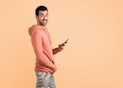 穿着粉红色运动衫的男人看着相机，微笑着在Ocher背景上使用手机