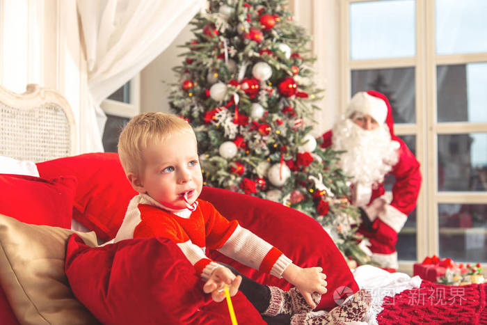 小悲伤的男孩坐在节日卧室的床上。 真正的圣诞老人躲在圣诞树后面。