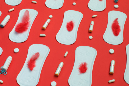 红色羽毛和白色垫堆许多卫生棉条和药丸在红色背景。月经, 卫生保护手段
