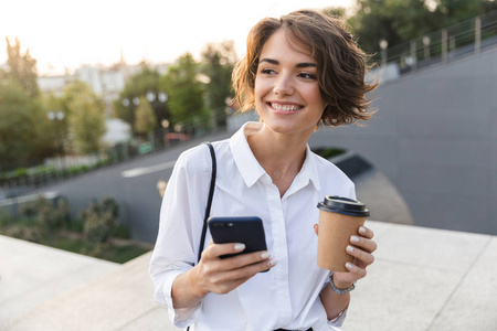 微笑的年轻女子站在街上，用手机端着杯咖啡