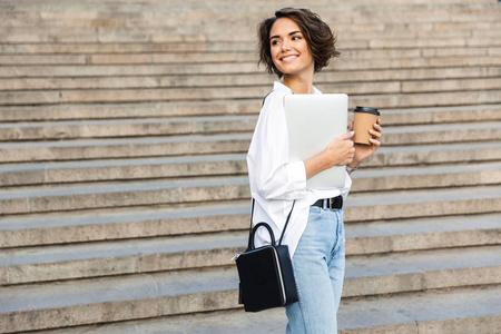 年轻可爱的漂亮女人在户外散步的照片，手提电脑拿着咖啡。