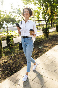 可爱的年轻女子拿着手机在街上散步，喝着咖啡