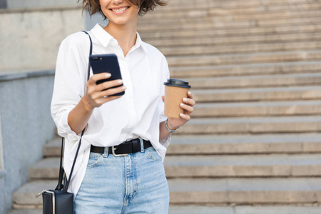 裁剪照片的年轻惊人的女人在街上使用手机拿咖啡。