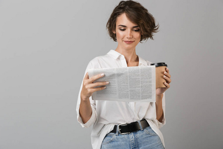 照片上一个快乐的年轻女人摆出孤立的灰色背景，喝咖啡，拿着报纸看书。