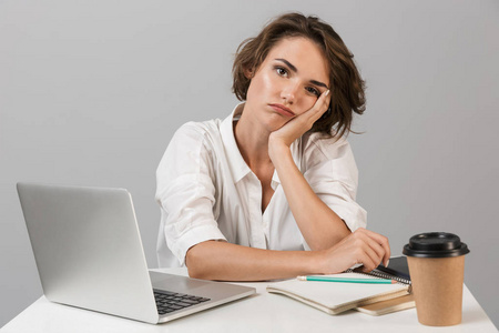一个无聊疲惫的年轻女人坐在桌子旁的照片，在笔记本电脑附近的灰色背景上被隔离。