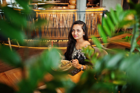 漂亮的印度女孩穿着黑色的裙，坐在餐厅里，端着果汁和沙拉。