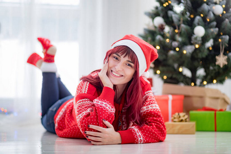 戴着圣诞帽的美丽的自然女人躺在圣诞树旁边的地板上，并赠送礼物
