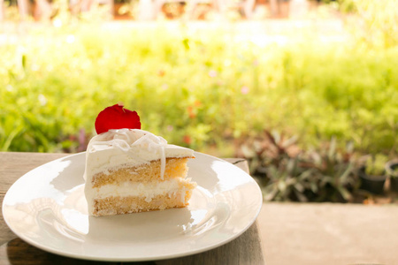 一块美味的椰子蛋糕，用红色玫瑰花瓣装饰在白色的盘子里，放在有绿色花园背景的木桌上。