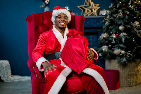 快乐微笑的非洲圣诞老人在他的房子旁边的壁炉和圣诞树休息在扶手椅。圣诞节假期