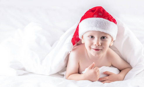 可爱的小男孩，戴着圣诞老人帽，躺在白色羽绒被下面的床上。