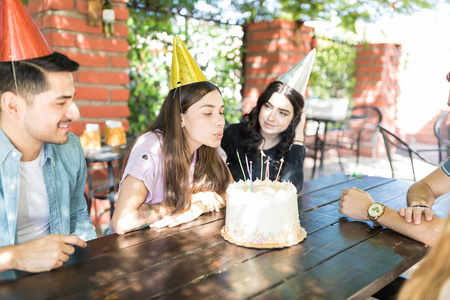 年轻的西班牙裔妇女在朋友包围的蛋糕上吹生日蜡烛