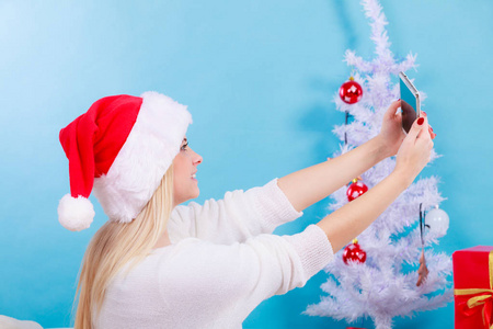 享受圣诞礼物。 戴圣诞帽的女人用智能手机拍照。 蓝色背景室内拍摄
