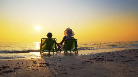 母亲和儿子戴着帽子坐在沙滩椅子上，看着夕阳溅满了海浪。 旅行概念