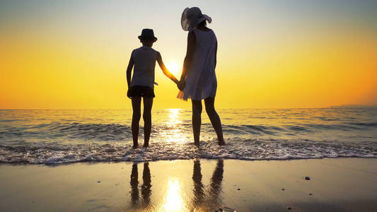 一家人在海滩上散步，到海边日落，溅起浪花。女人穿白色连衣裙和草帽，男孩戴帽子