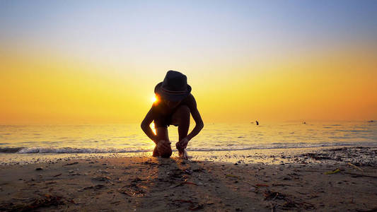 带帽子的男孩的剪影，扔石头在海水表面跳跃。 暑假概念，充满活力的橙色天空