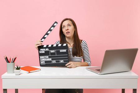 疲惫的女人滚动的眼睛，拿着经典的黑色电影制作剪贴板，并在项目上工作，而坐在办公室与笔记本电脑隔离在粉红色的背景。 成就事业。 复