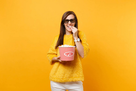 微笑的年轻女孩的肖像，在3DIMA X眼镜，看电影电影，吃爆米花从桶隔离在明亮的黄色背景。 人们真诚的情感在电影生活方式的概念。