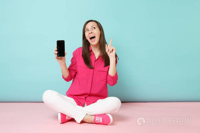 全长肖像年轻有趣的女人穿玫瑰衬衫，白色裤子，坐在地板上，手机隔离在明亮的粉红色蓝色粉彩墙背景工作室。 时尚生活方式的概念。 模拟