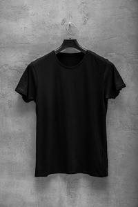 男黑色棉T衬衫的正面在衣架上，背景上有一堵混凝土墙。 T恤没有印花