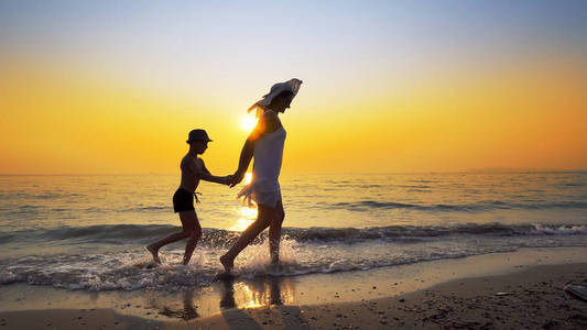 暑假的家人。 母亲穿着白色连衣裙，儿子戴着帽子，在生机勃勃的夕阳下，漫步在宁静的海滩上