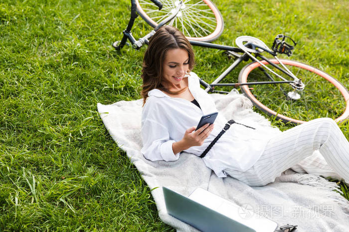 骑自行车的漂亮年轻女子坐在草地上，手提电脑拿着手机