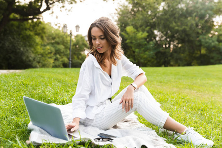 微笑的年轻女子在公园里坐在草地上，用笔记本电脑