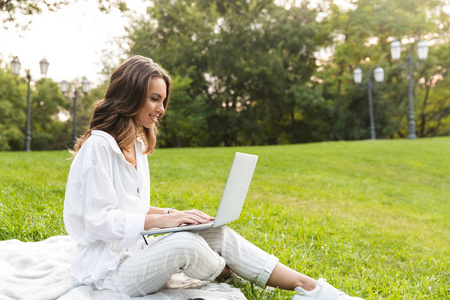 快乐的年轻女子在公园里坐在草地上，用笔记本电脑