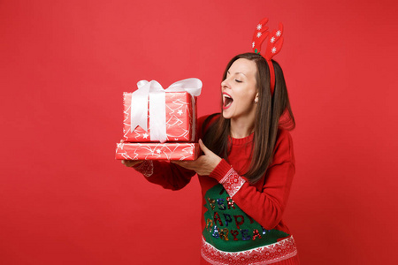 欢快的年轻圣诞老人女孩在有趣的装饰鹿角，拿着红色的盒子和礼物，保持嘴巴张开，孤立在红色的背景上。 新年快乐2019庆祝节日派对概